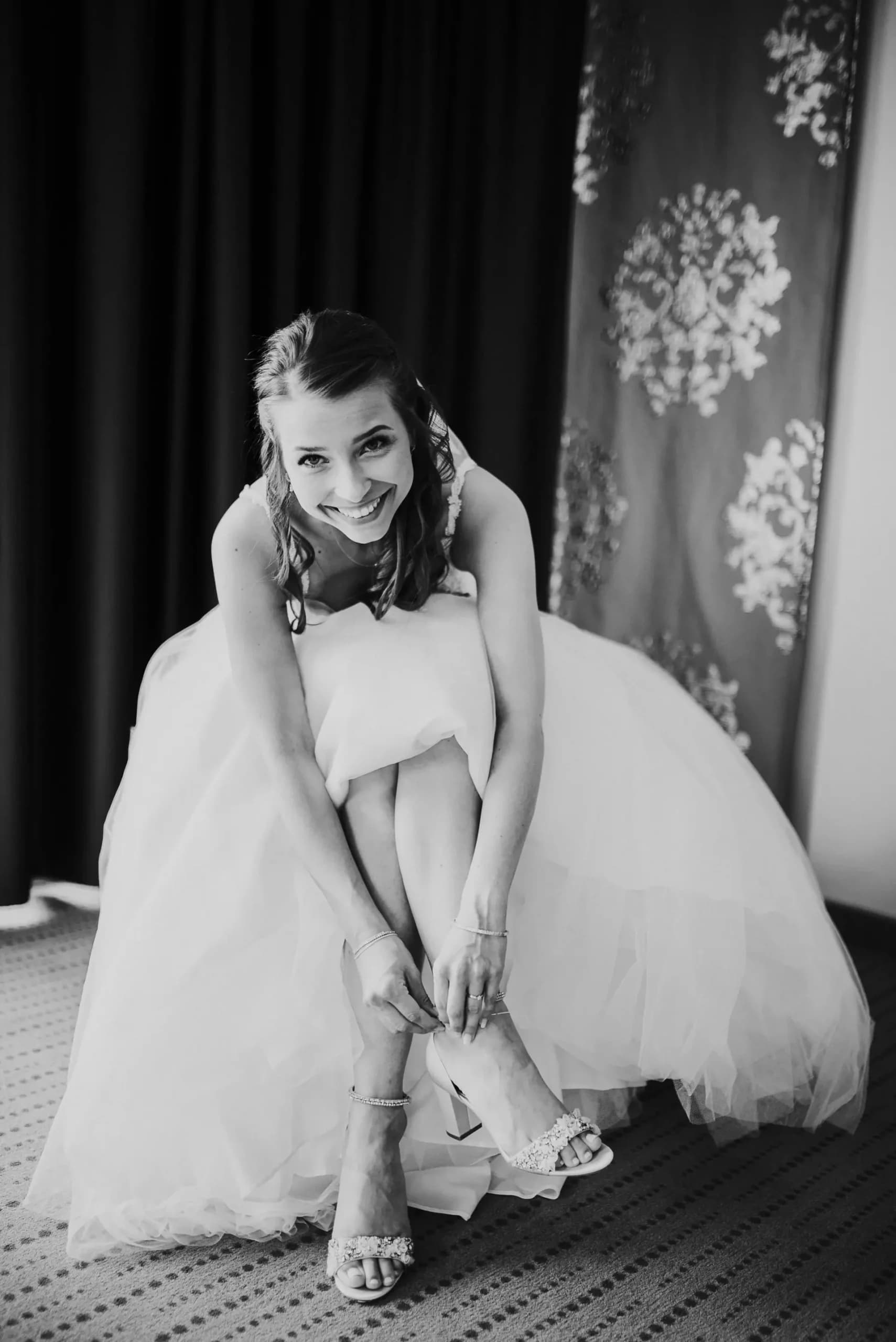 Braut in weißem Hochzeitskleid sitzt lächelnd und zieht ihre Schuhe an
