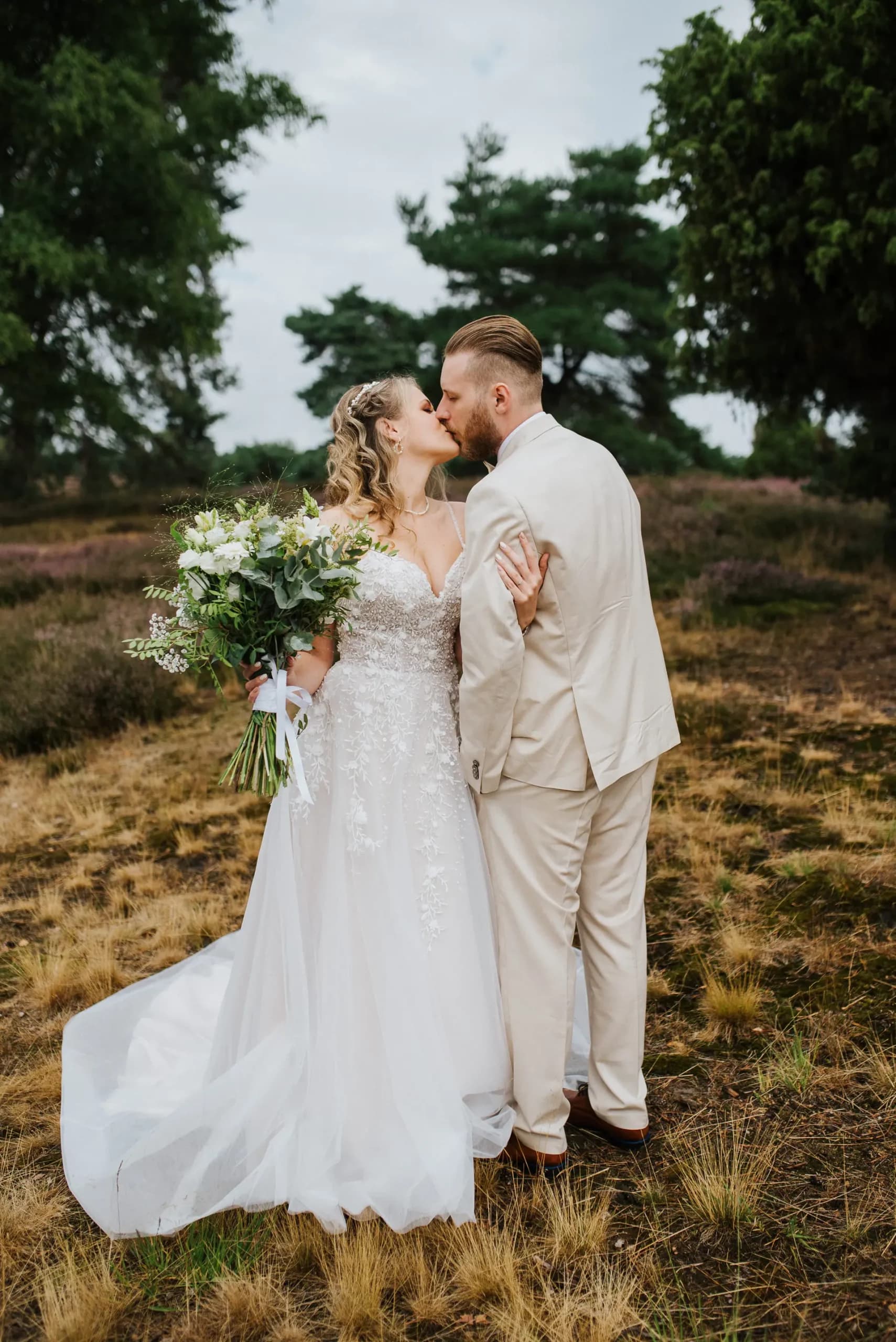 Ein Brautpaar küsst sich in einem ländlichen Feld mit Bäumen im Hintergrund, die Braut trägt ein weißes Kleid und hält einen Blumenstrauß