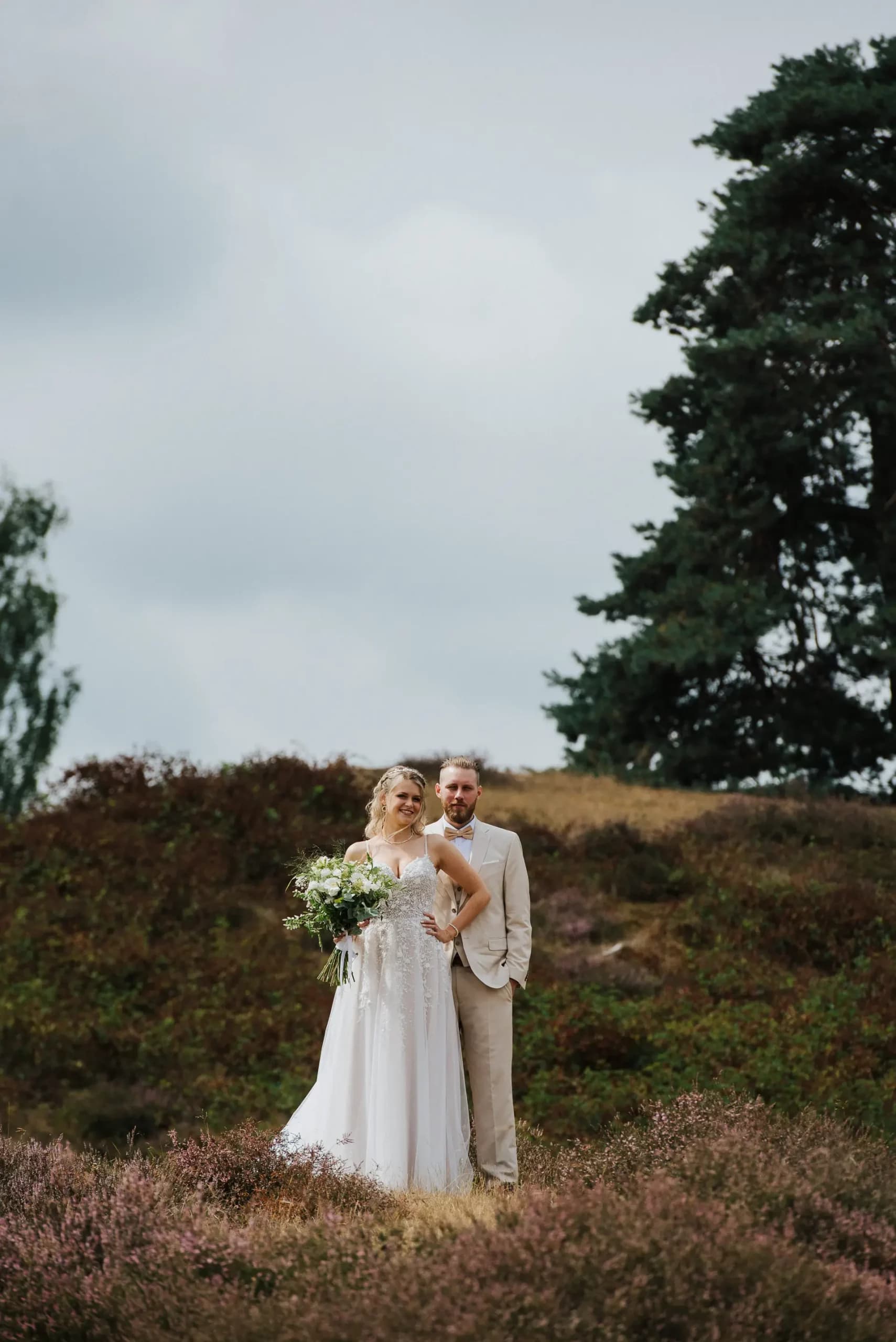 Brautpaar in Hochzeitskleidung posiert in blühender Heideauch mit Wildblumen-Strauß vor bewölktem Himmel